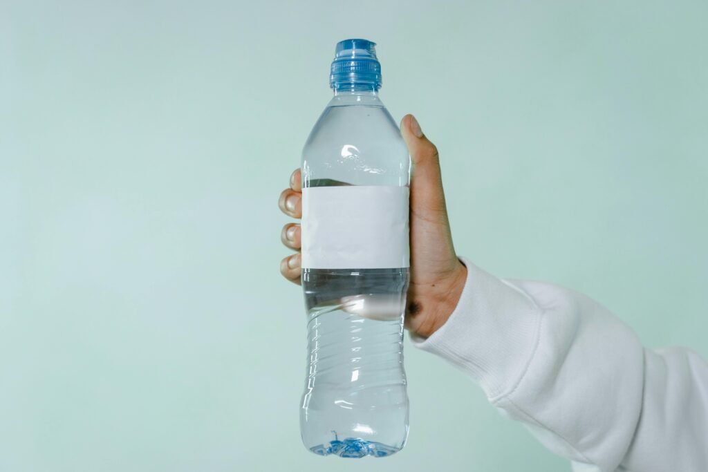 A hand holding a clear plastic water bottle with blank label on a soft green background.
